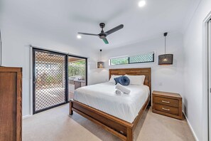 A bedroom with a wooden bed, a ceiling fan, a small bedside table, and a view through a window. - Whitsunday Sanctuary Retreat - Mountain Views (Jubilee Pocket)