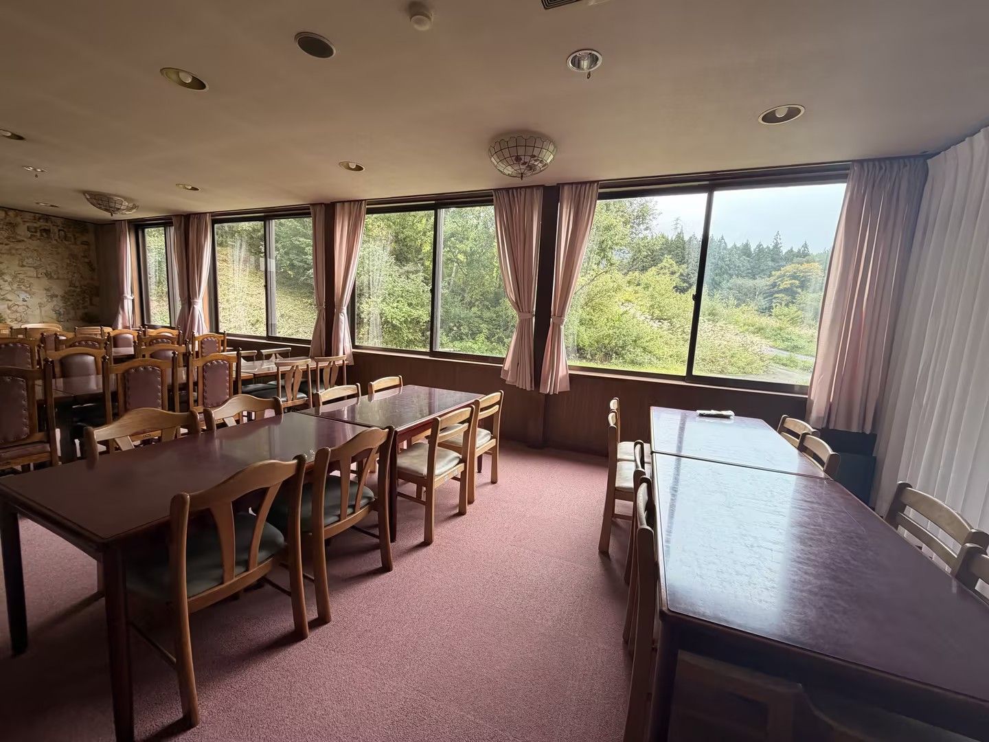A dining area with multiple wooden tables and chairs, large windows with curtains, and a stone wall.