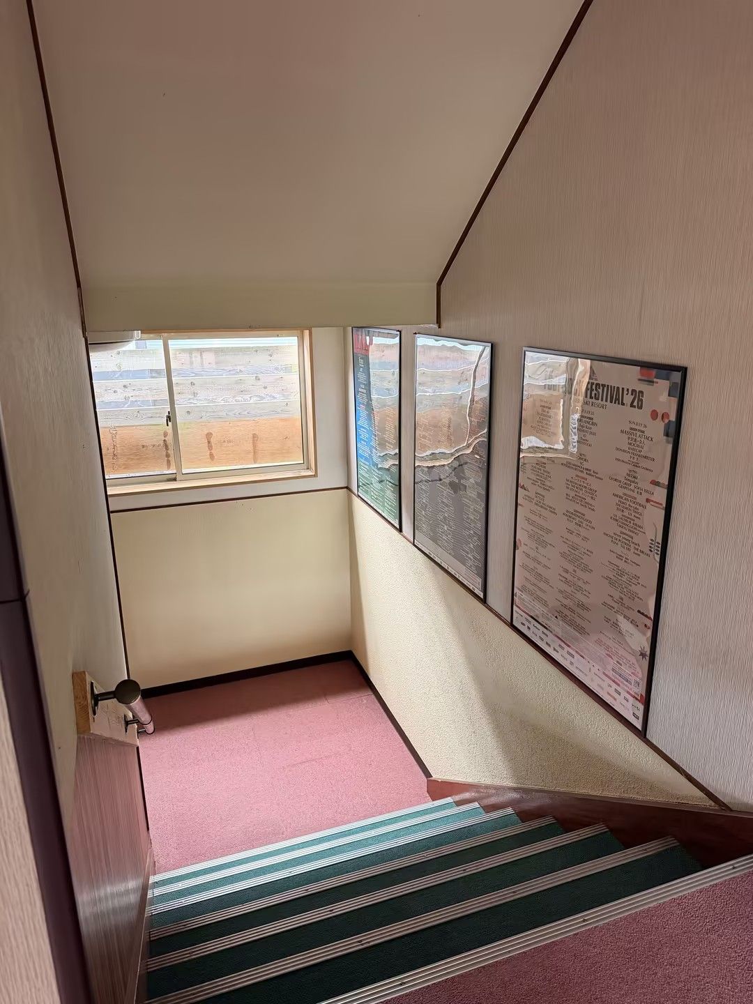 A corner room with a window, a poster on the wall, and a staircase with a striped carpet.