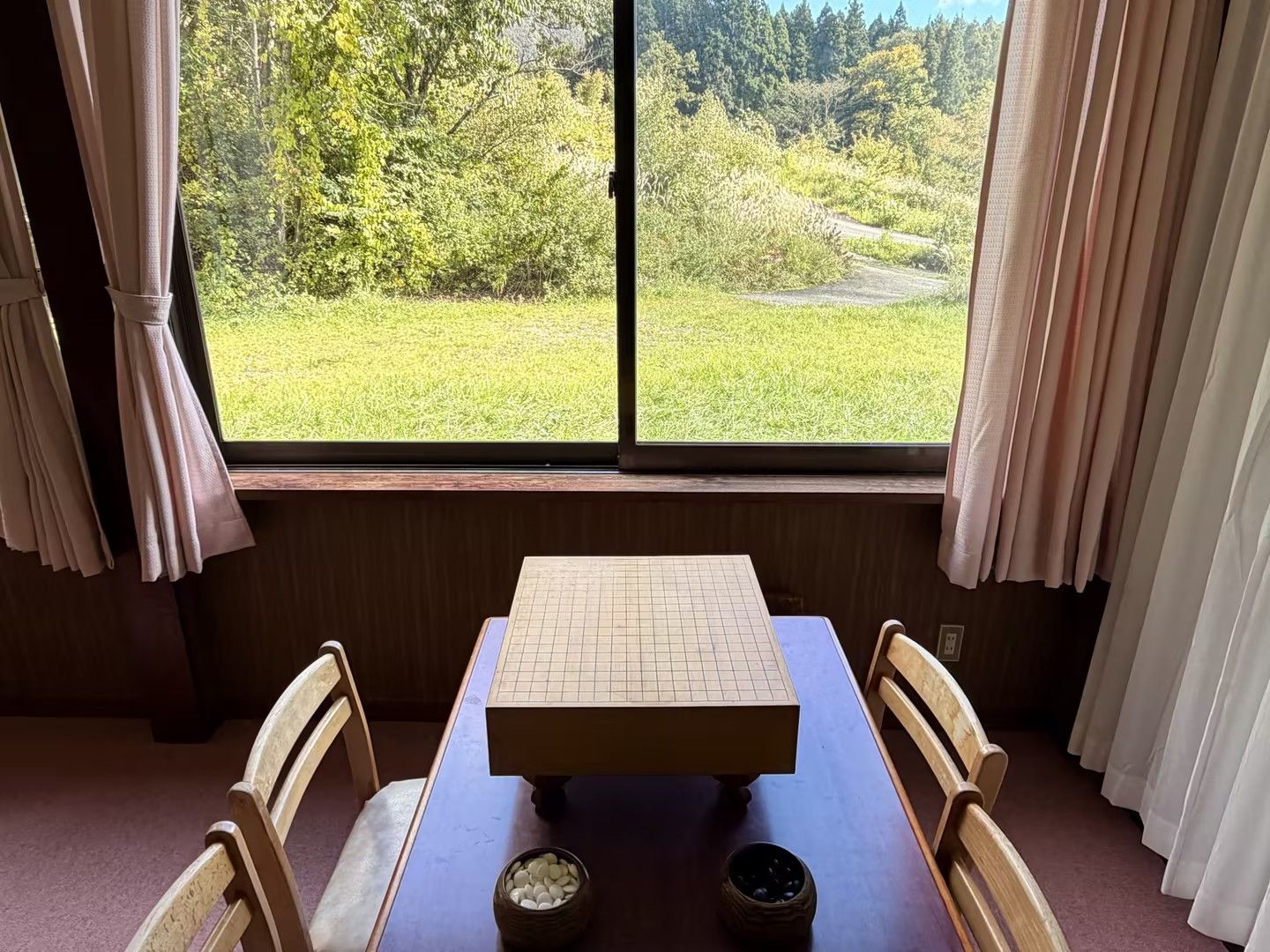 Ein Zimmer mit Tisch und Stühlen, einem fenster mit Vorhängen und Blick auf eine grüne Fläche.