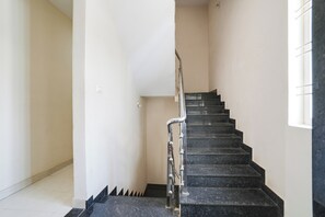 Una escalera con escalones negros y barandal de metal que lleva a un rellano con pared blanca y un interruptor de luz.