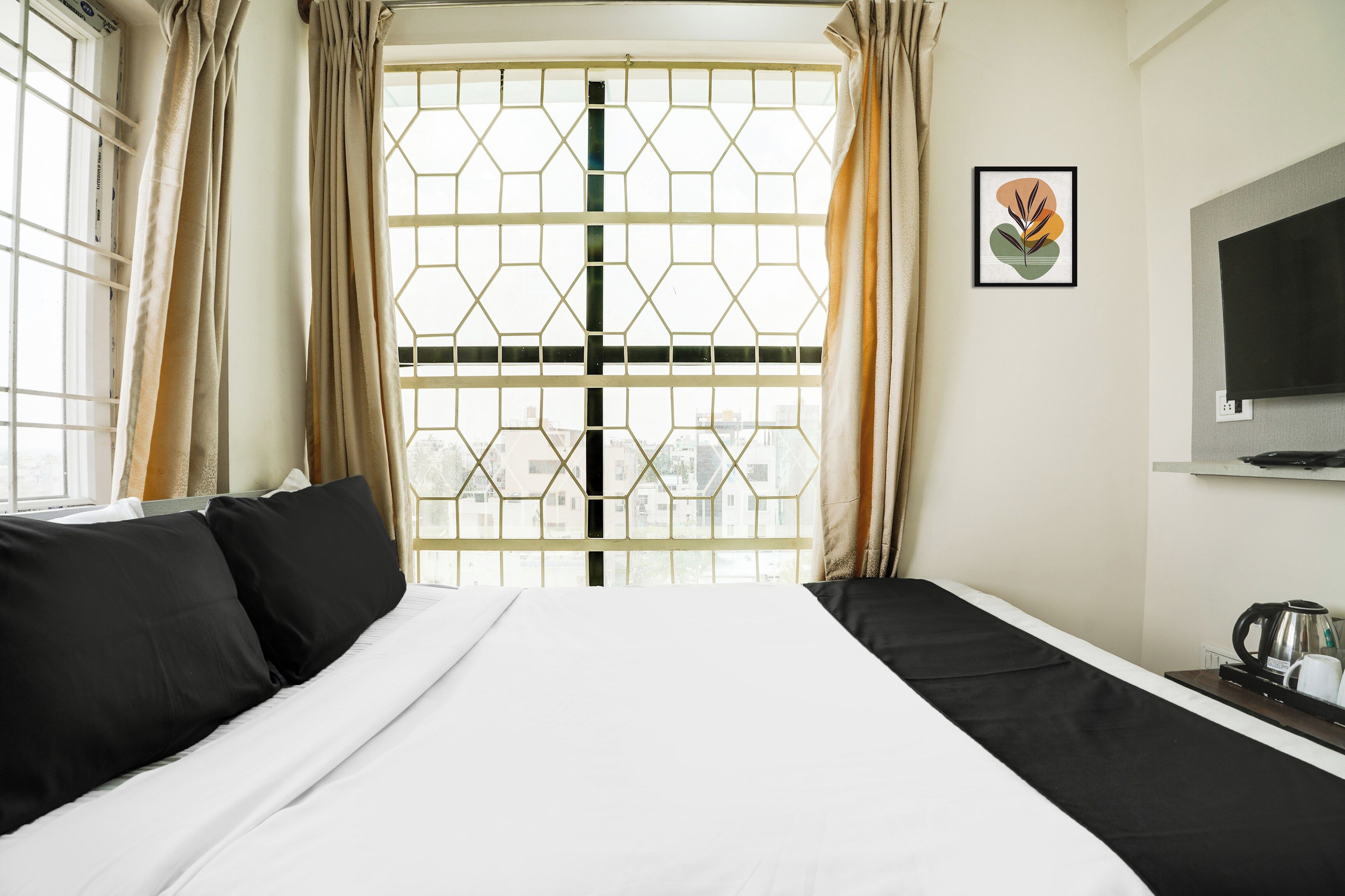 Una habitación de hotel con un ventanal grande, una cama con ropa de cama blanca y negra, un televisor montado en la pared y una tetera sobre una mesa.