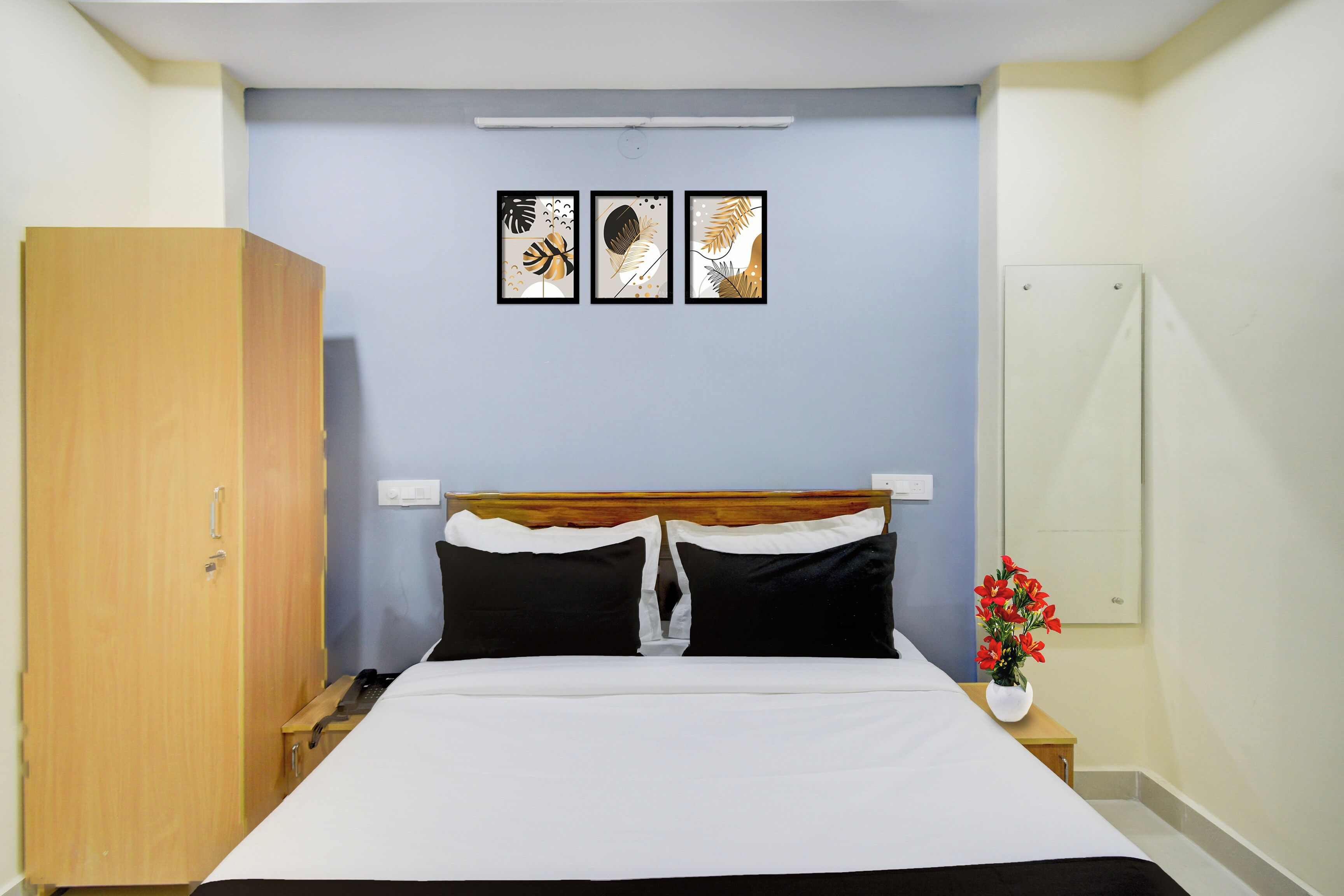 Una habitación de hotel con una cama, un armario, una mesita de noche y una mesita auxiliar con un jarrón de flores, además de tres cuadros decorativos en la pared.