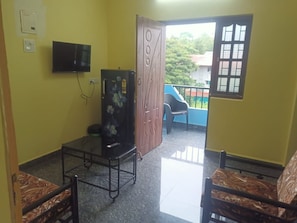 A room with a television, a wooden door, a window, and a balcony with a blue railing.