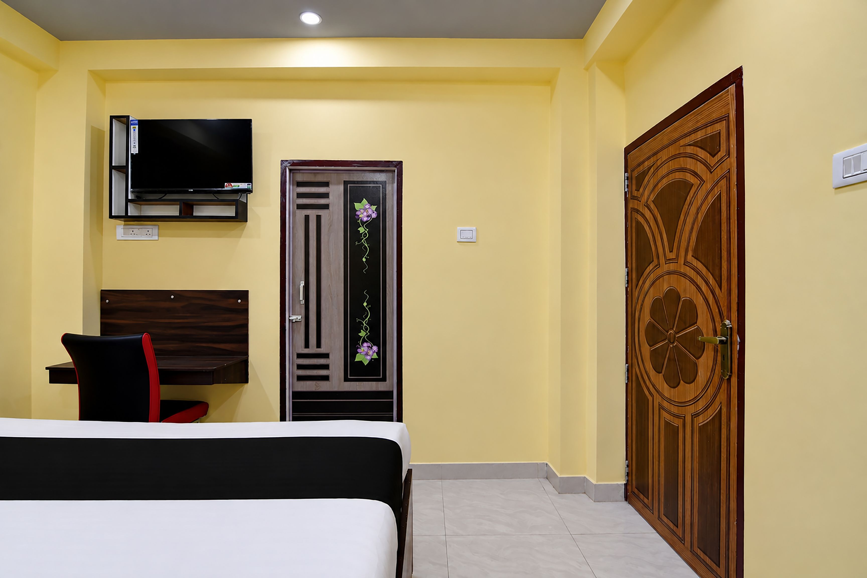 Ein Hotelzimmer mit Bett, Schreibtisch, Fernseher, einer Tür mit dekorativer Verkleidung und einem Lichtschalter an der Wand.