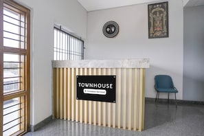 Un mostrador de recepción con un letrero que indica servicio de la empresa, una ventana con rejas, una silla azul y un icono religioso enmarcado en la pared.