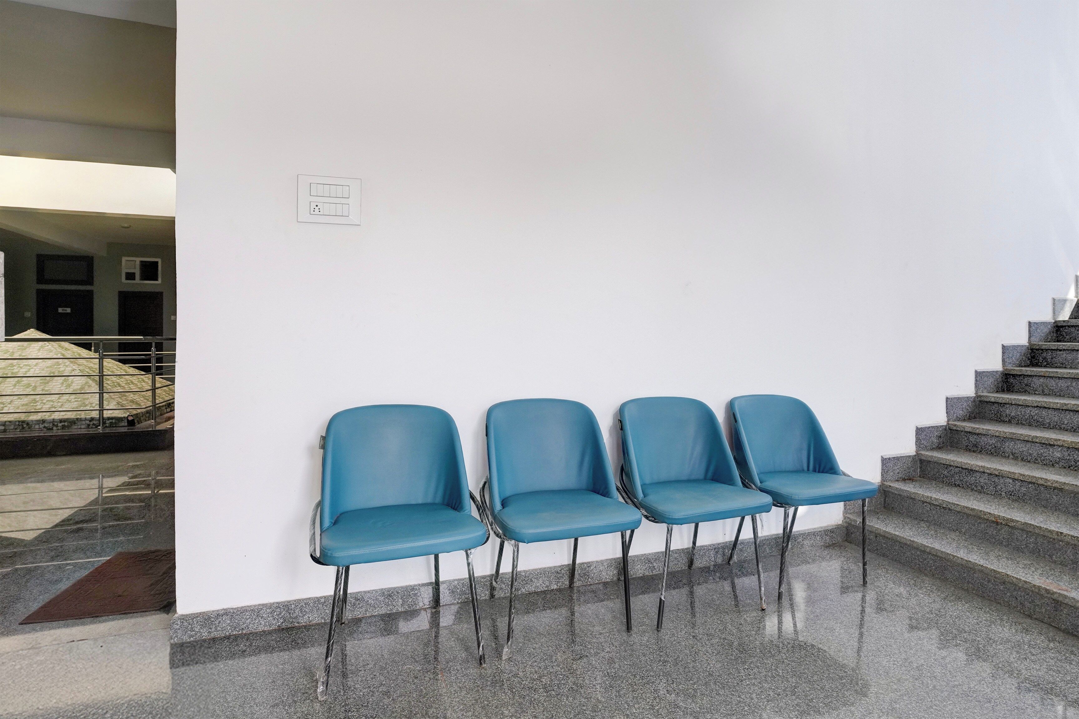 A set of four blue chairs against a white wall, positioned on a polished stone floor with a staircase to the right.