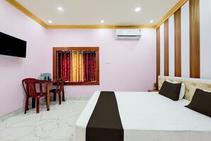 A hotel room with a bed, a desk with chairs, a window with curtains, an air conditioning unit, and a television mounted on the wall.