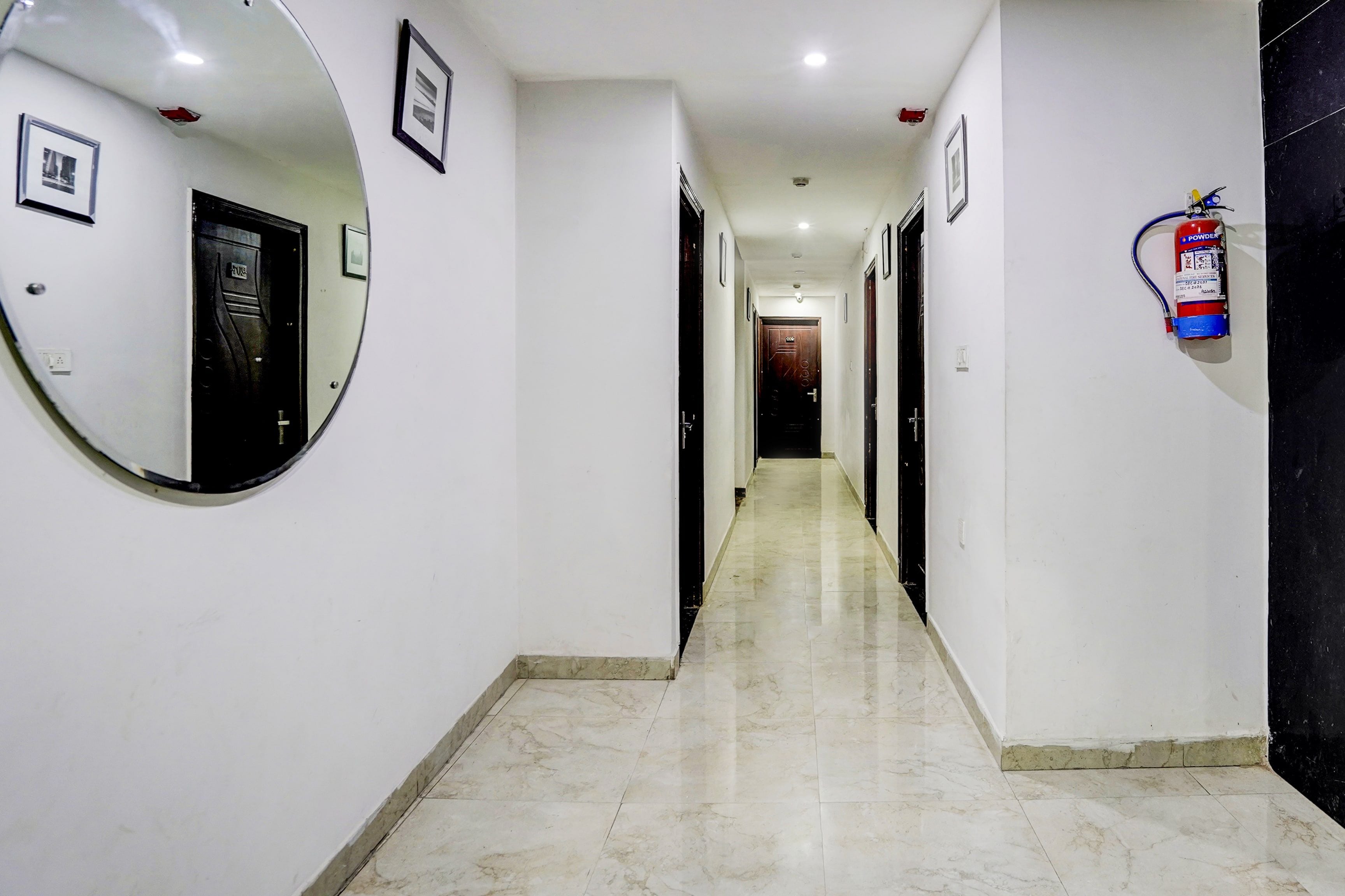 A corridor with a round mirror, framed pictures, a fire extinguisher, and a door at the end.