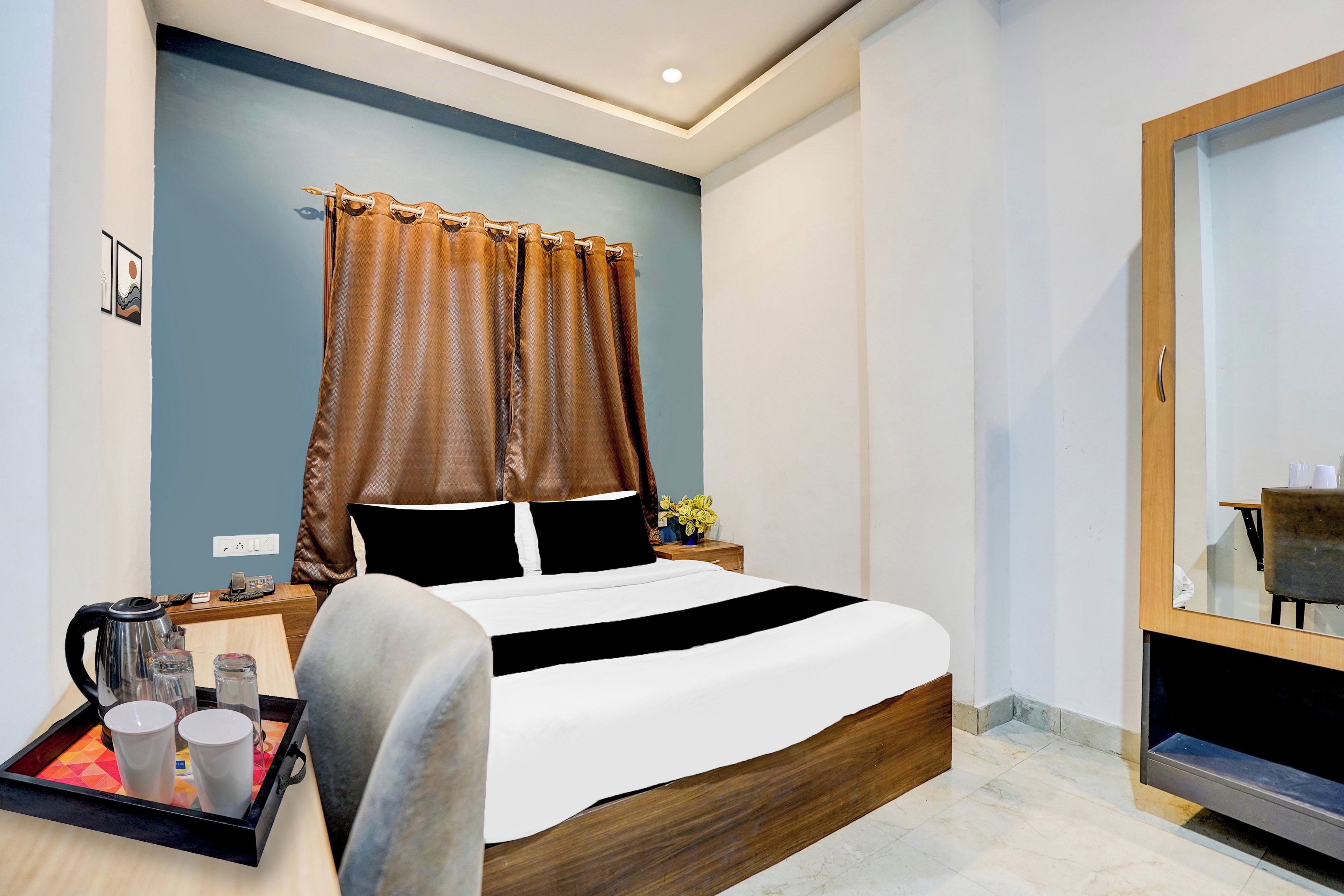 A hotel room with a bed, a bedside table with a kettle and cups, a mirror, and a tray with cups.