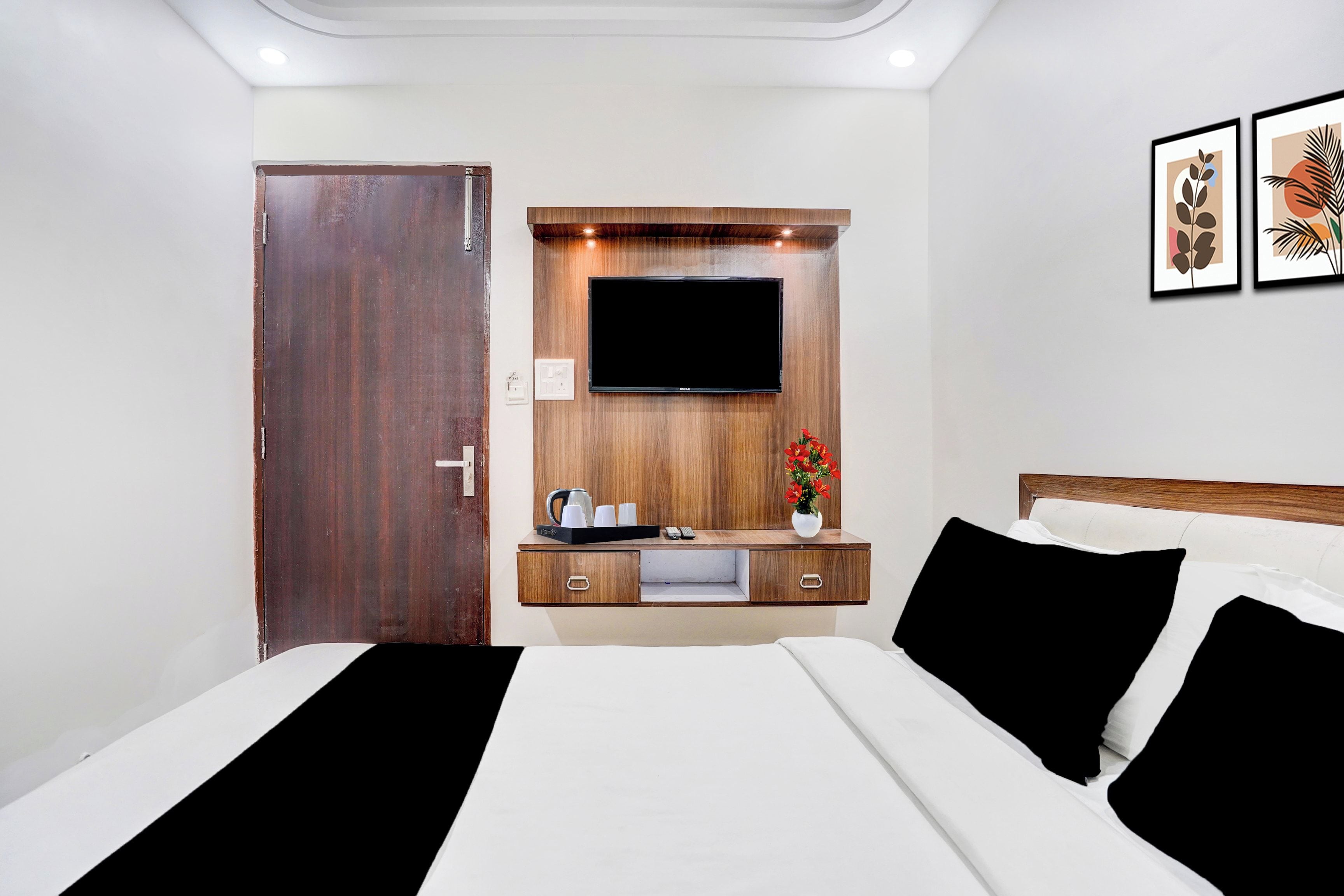 A hotel room with a bed, a TV mounted on a wooden panel, a small table with a vase of flowers, and a door.