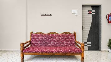 A wooden sofa with a floral pattern, a tiled floor, a door with a checkered pattern, and a fire extinguisher mounted on the wall.