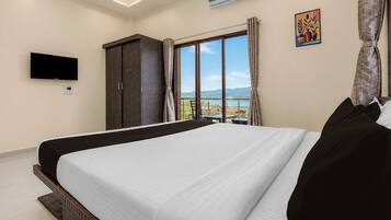 A hotel room with a large bed, a flat-screen TV, an air conditioner, a window with a view of the mountains, and a painting on the wall.