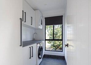 A compact laundry room with a washing machine, a sink, and a window providing a view of greenery outside. - Private Creek | Executive Home | Kayaks & Zipline (Sunshine Coast)