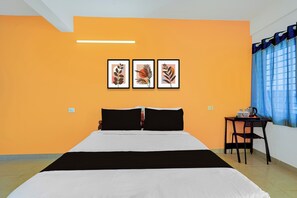 A hotel room with a bed, a small table with a kettle, and three framed artworks on the wall.