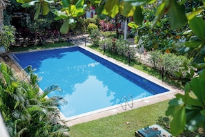 A rectangular swimming pool with clear blue water, surrounded by a well-maintained lawn and various tropical plants. - Charming 3-bedroom apartment with AC in peaceful Candolim (Arpora)