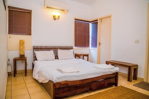 A bedroom with a wooden bed, a bedside table, a nightstand, a wall-mounted light fixture, and a window with blinds. - Charming 3-bedroom apartment with AC in peaceful Candolim (Arpora)