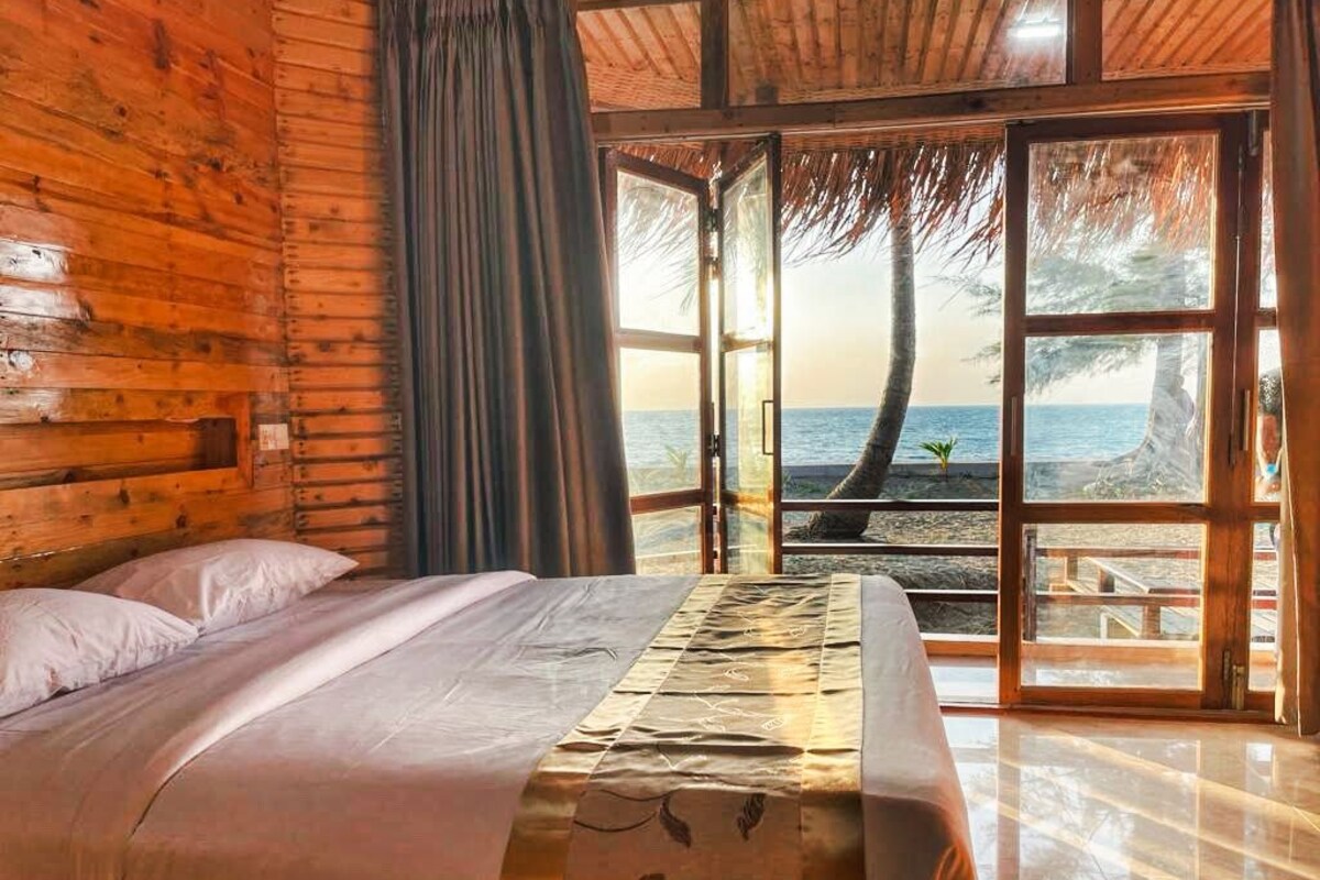 Habitació de fusta amb un llit, vista al mar i teulada de palla.