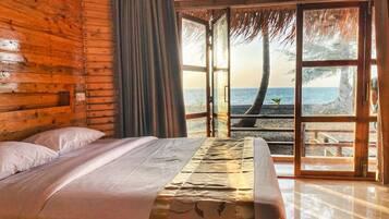 A wooden room with a bed, a view of the sea, and a thatched roof.