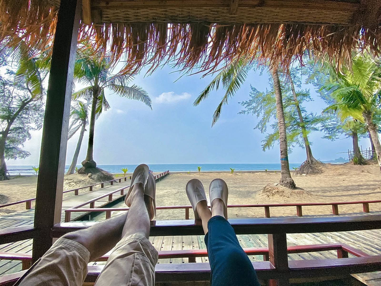 A wooden deck with a thatched roof overlooking a beach with palm trees and a clear sky.
