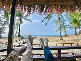 Um deck de madeira com telhado de palha com vista para uma praia com palmeiras e céu claro.