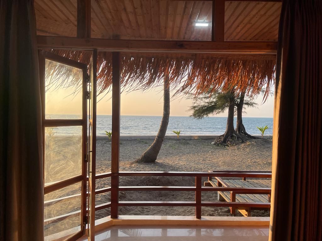 A beach view from a wooden-framed room with a thatched roof, a wooden deck, and a railing.
