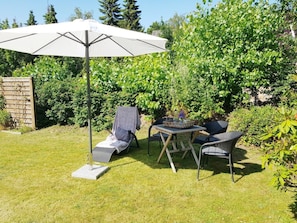 En utomhusplats med bord och stolar, ett stort parasoll och frodig grönska i bakgrunden.