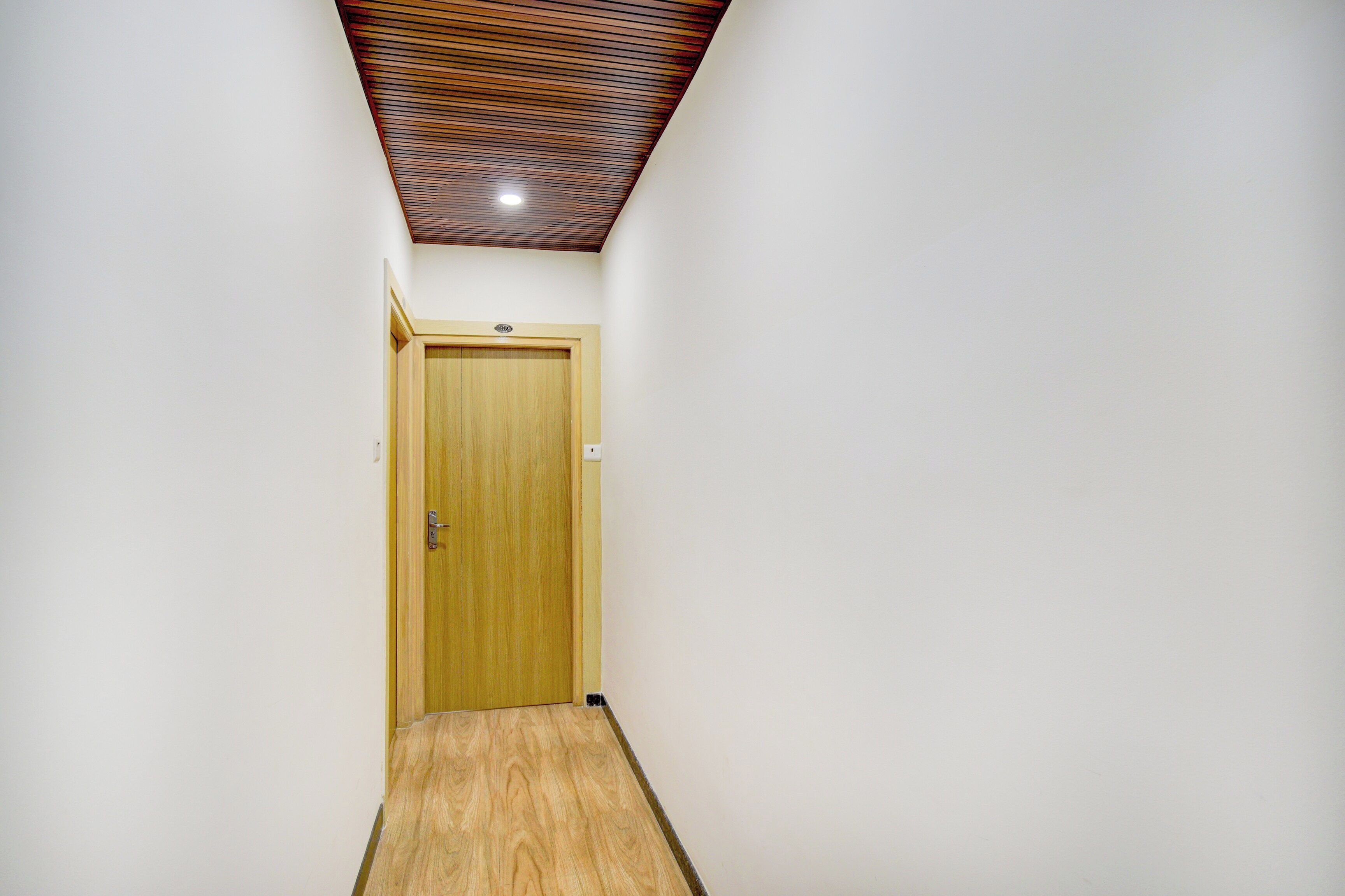 Un pasillo estrecho con una puerta de madera al final, techo inclinado y un interruptor de luz en la pared.