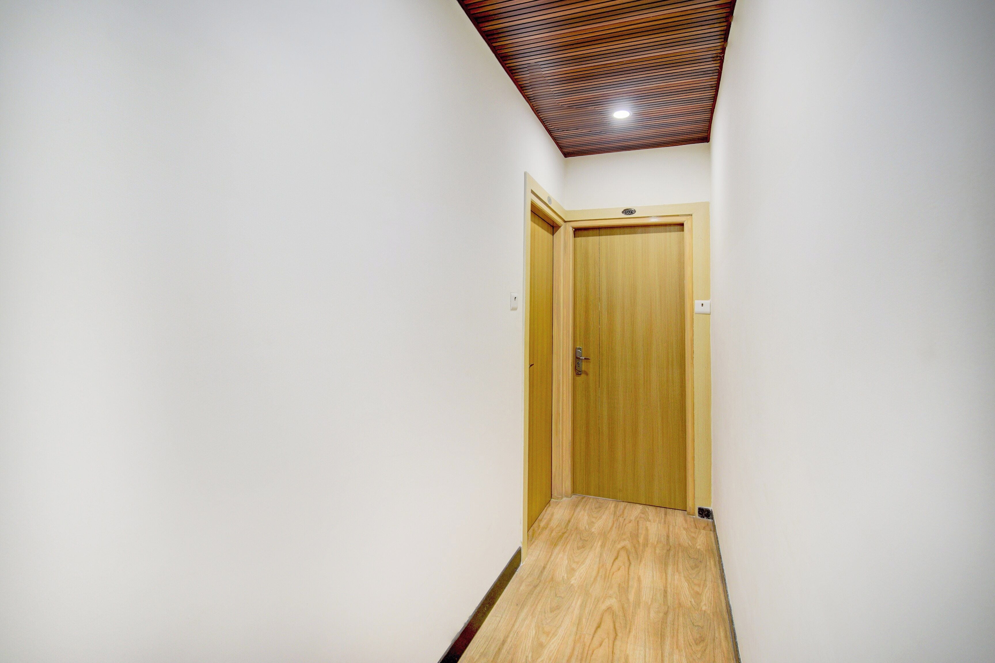 A narrow hallway with a wooden door at the end, a ceiling with a slatted design, and a light fixture.