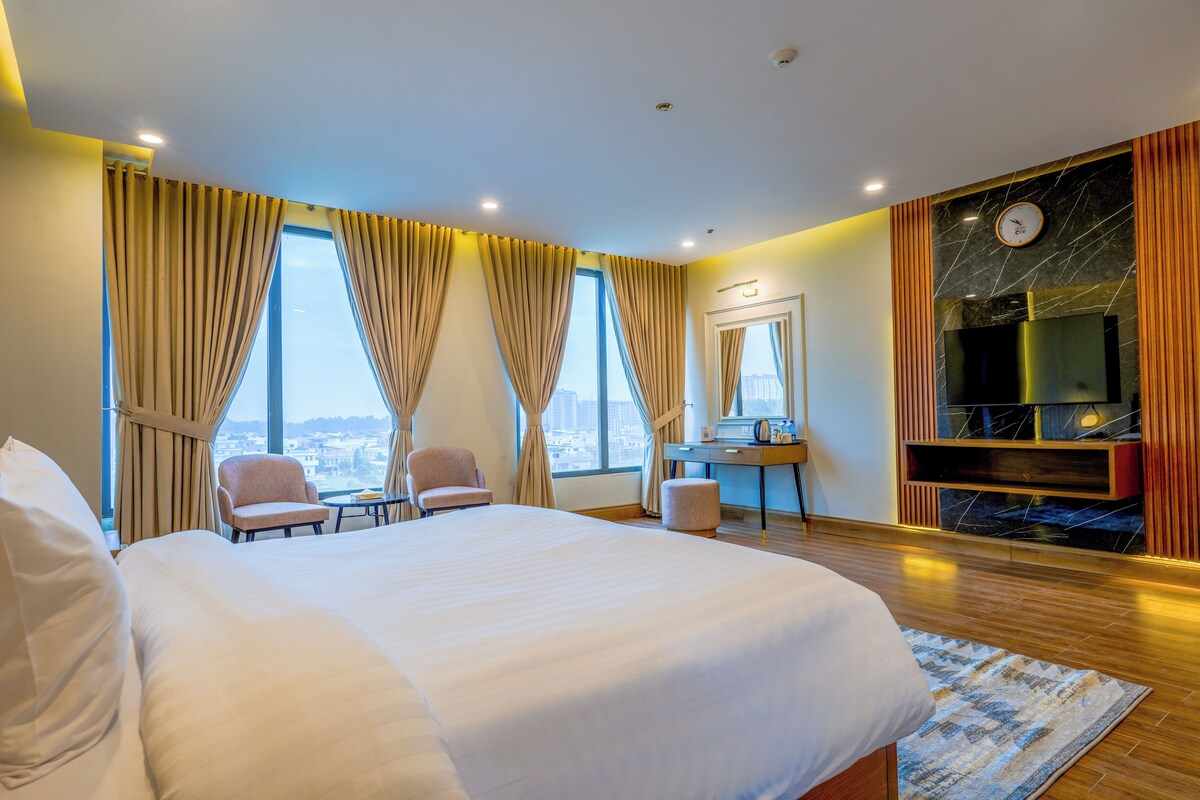 A hotel room with a large bed, two armchairs, a desk, a mirror, and a TV.
