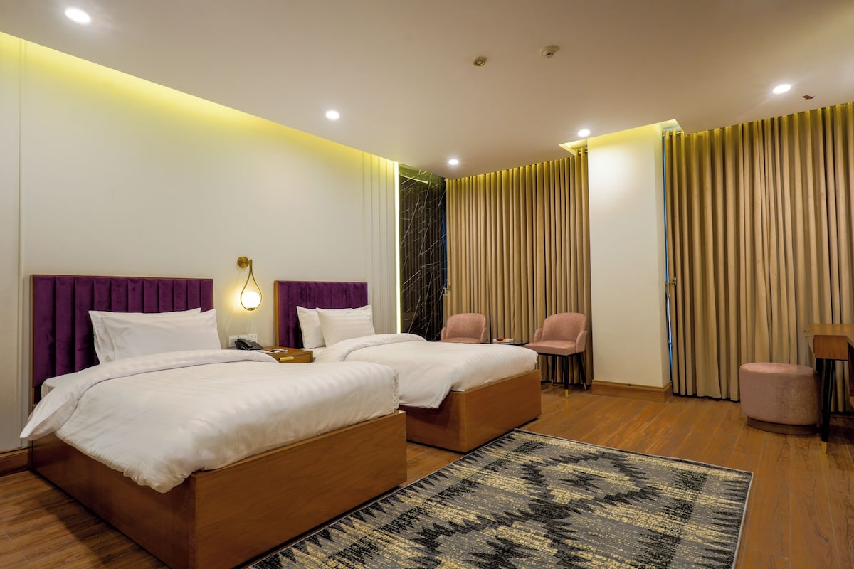 A hotel room with a double bed, two bedside tables, a desk, a chair, and a large window with curtains.