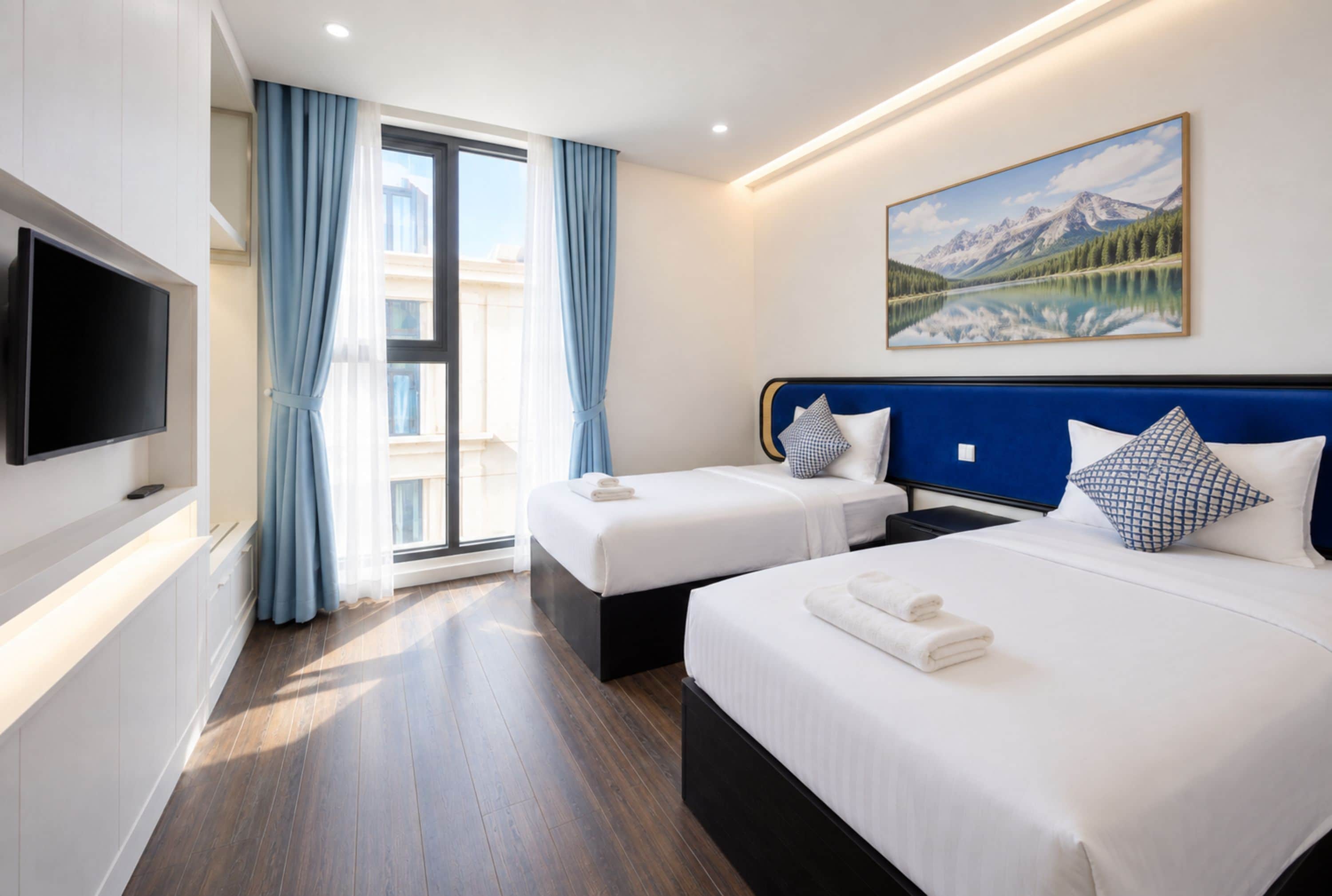 Une chambre d’hôtel avec deux lits, un téléviseur à écran plat, un tableau représentant un paysage de montagne et une grande fenêtre avec des rideaux bleus.