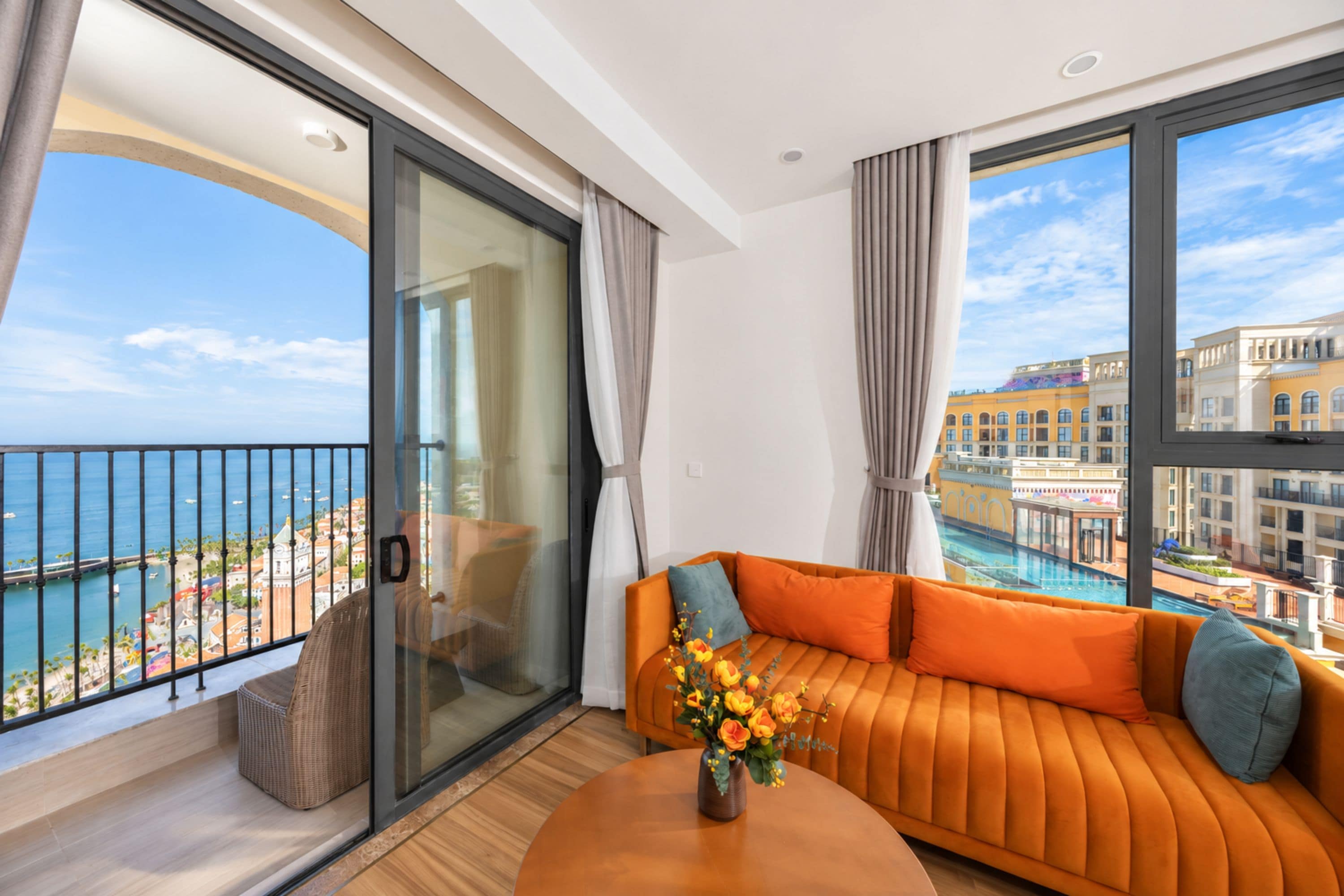 A modern hotel room with a balcony, a sofa, and a view of the sea and buildings.