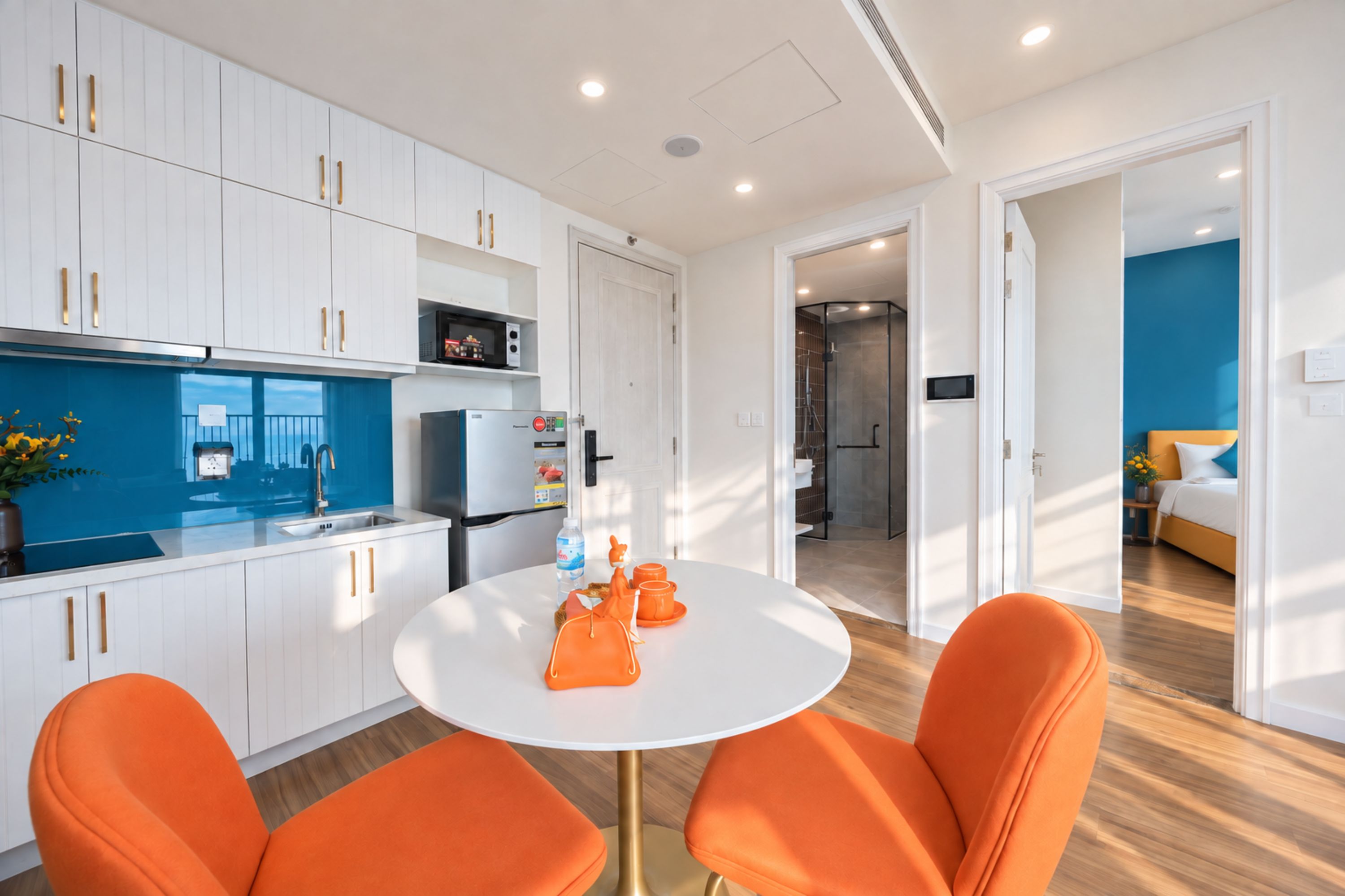 A modern kitchen with white cabinets, a refrigerator, and a microwave. There is a round table with two orange chairs.