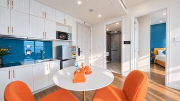 A modern kitchen with white cabinets, a refrigerator, and a microwave. There is a round table with two orange chairs.