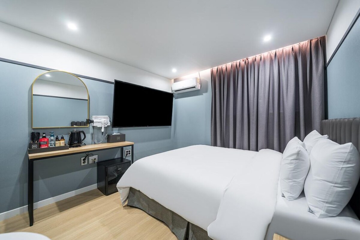 A modern hotel room with a bed, a desk with a flat-screen TV, and a large mirror.