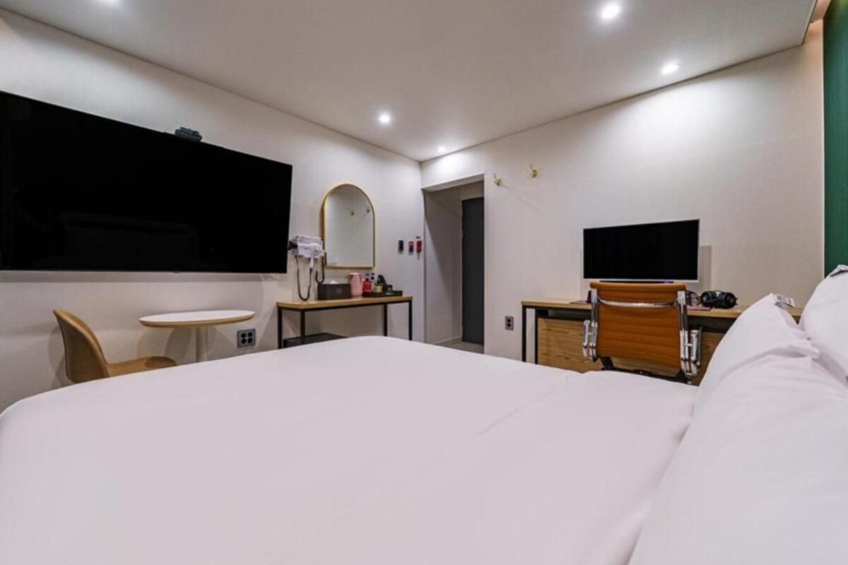 A hotel room with a large bed, a desk, a chair, a small table, a mirror, and a television.