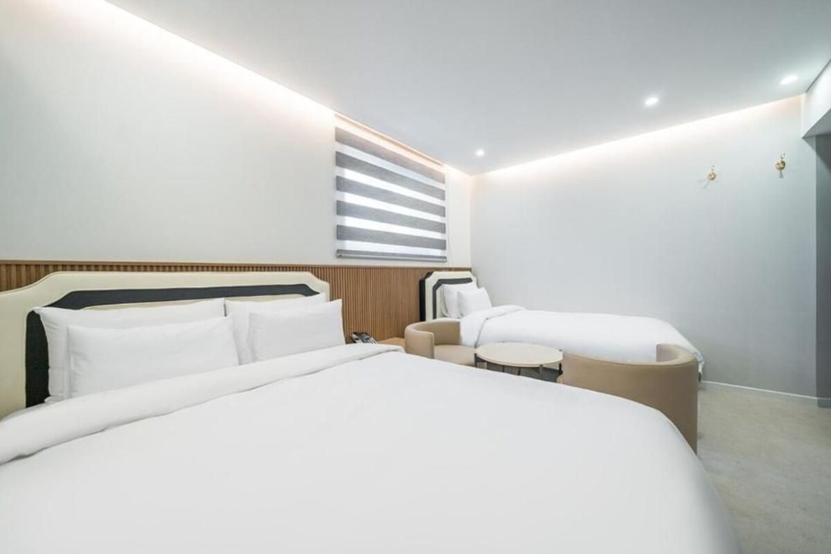 A modern hotel room with a large bed, a smaller sofa, a round table, and a window with blinds.