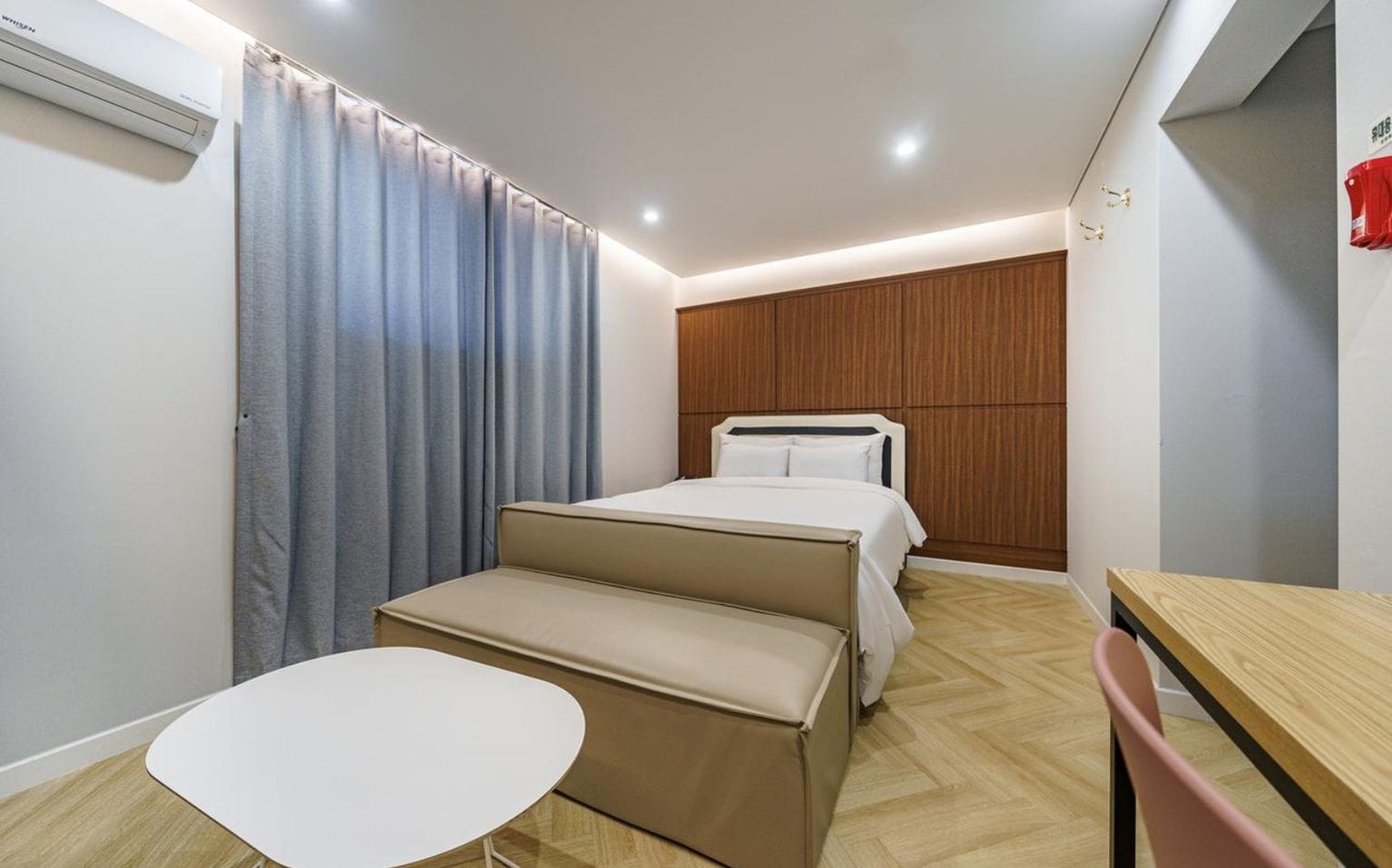 A compact hotel room with a bed, a small table, a bench, and a wooden wardrobe.