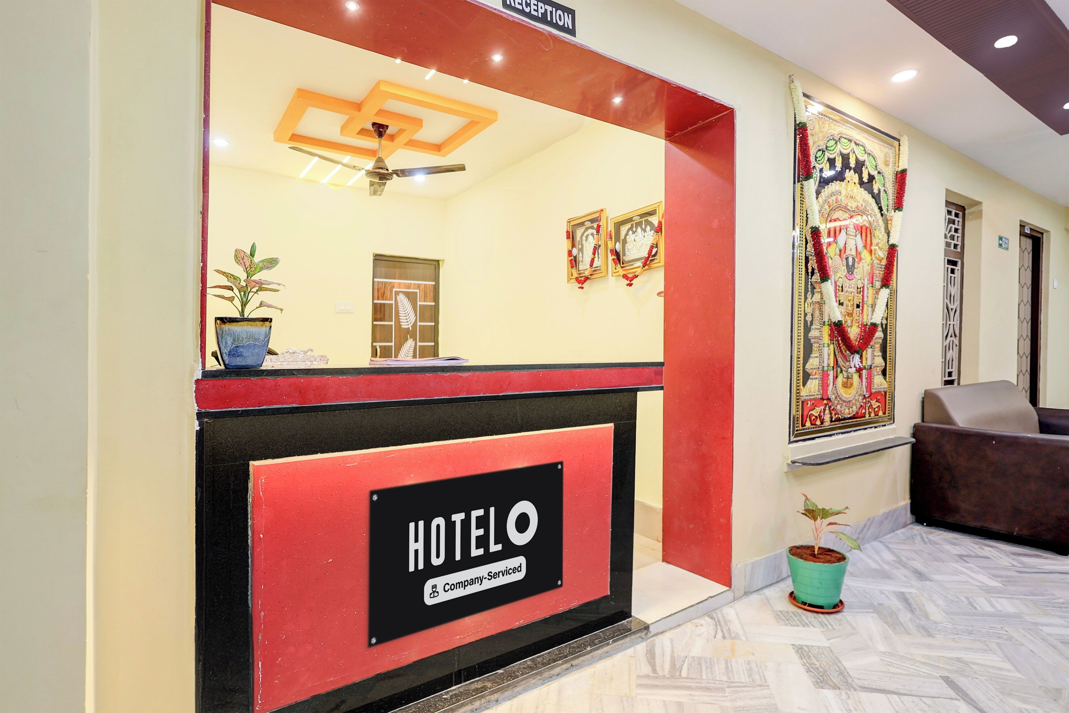 A hotel reception area with a reception desk, a ceiling fan, and a wall-mounted sign indicating the hotel name.