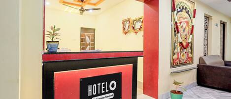 A hotel reception area with a reception desk, a ceiling fan, and a wall-mounted sign indicating the hotel name.