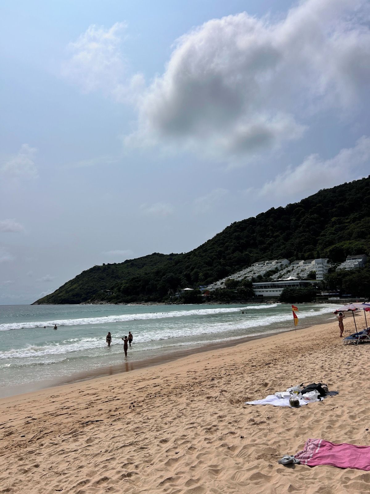 Ein Strand mit Menschen, die am Ufer entlanggehen, feiner Sand und ein Hügel mit Gebäuden im Hintergrund.