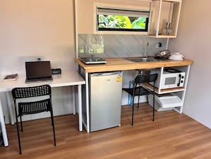 Eine kompakte Küchenzeile mit integriertem Mikrowellenherd, einem kleinen Kühlschrank und einem Spülbecken. Auf einem weißen Schreibtisch steht ein Laptop, und es gibt zwei schwarze Stühle.