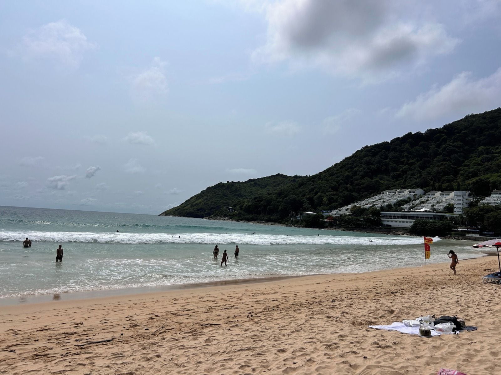 Une plage où l’on voit des gens nager et se promener, un rivage sablonneux et, au loin, une colline parsemée de bâtiments.