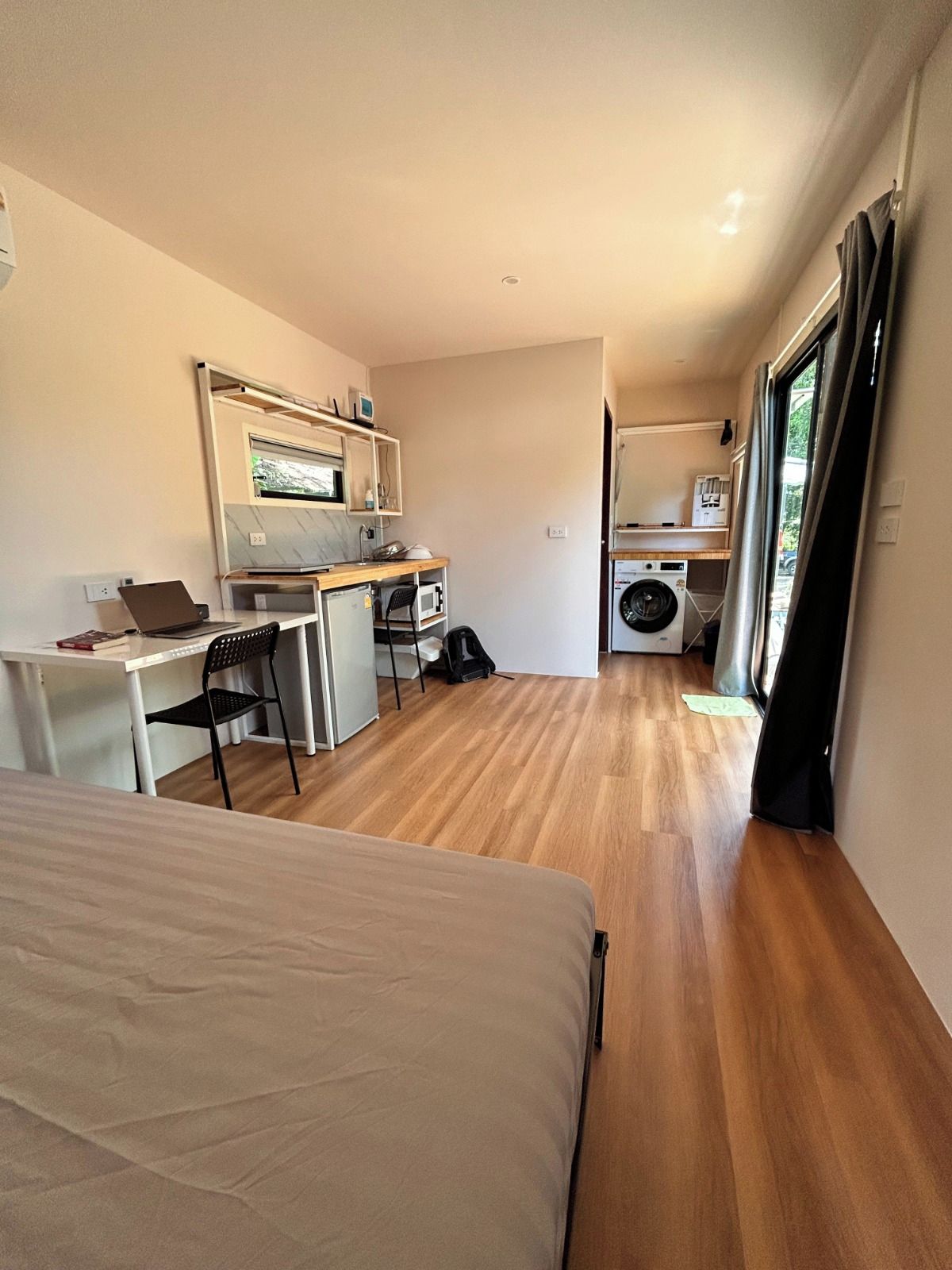 Ein kleines Zimmer mit einem Bett, einem Schreibtisch, einem Stuhl, einer Mikrowelle, einer Waschmaschine und einem Fenster.