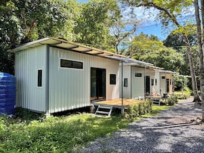 Fileira de casas móveis térreas com varandas, cercada por árvores e um caminho de cascalho.