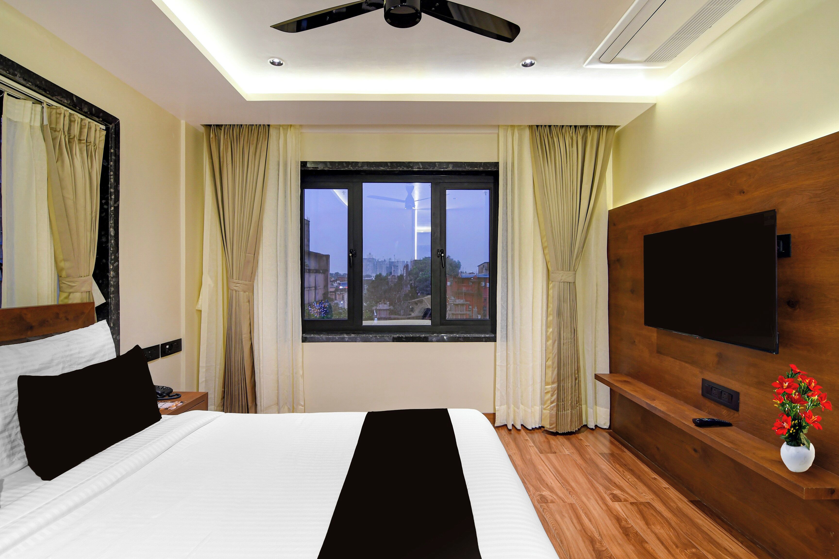 A hotel room with a bed, a TV, a wooden headboard, a window with curtains, and a ceiling fan.