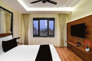 Habitación de hotel con cama, televisor, cabecero de madera, ventana con cortinas y ventilador de techo.