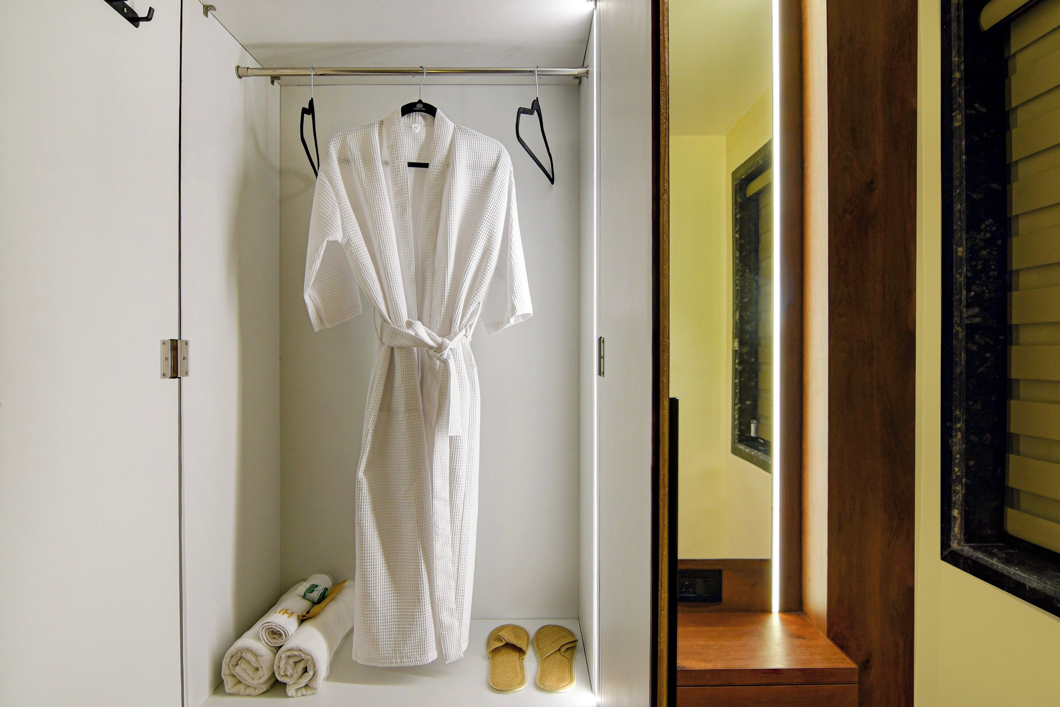 A white robe hanging on a metal rod in a bathroom with a wooden bench and a pair of slippers.