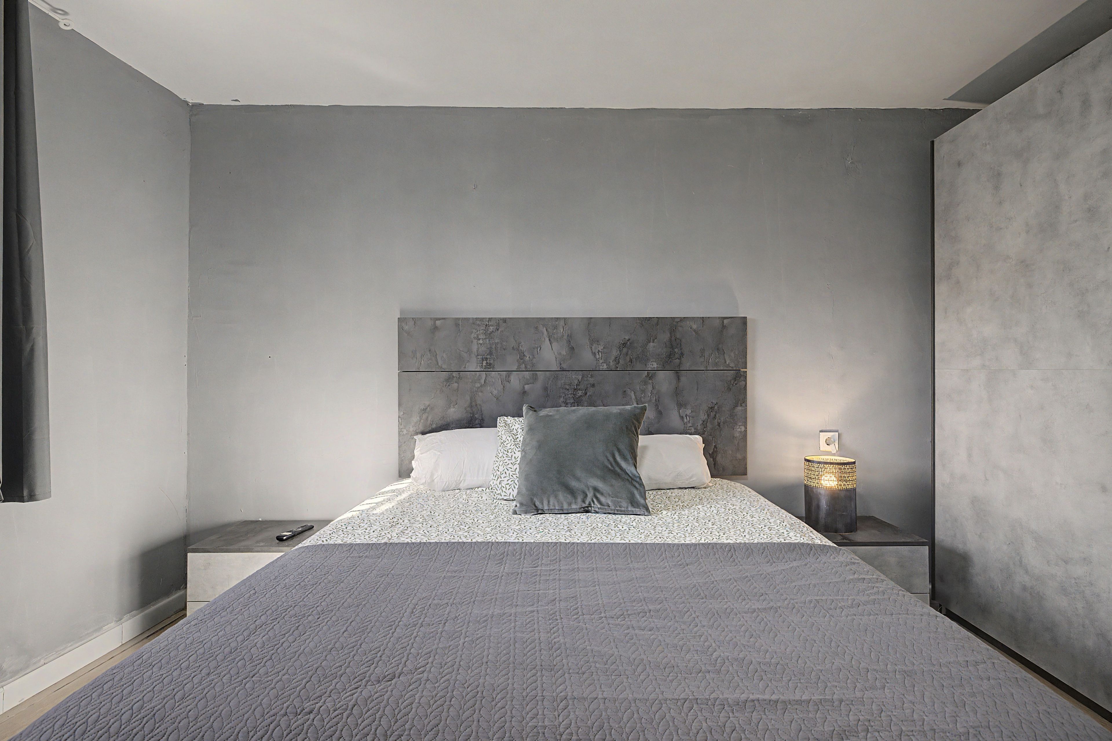 Een slaapkamer met een groot bed, een nachtkastje, een lamp en een grijs hoofdeinde.